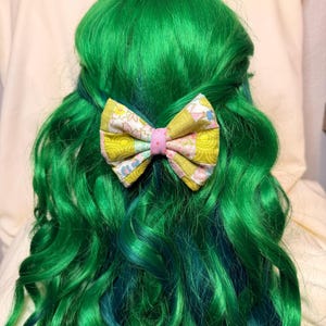 May include: A fabric hair bow featuring a patchwork design of yellow, pink, and white floral and geometric patterns. The bow is centred with a solid pink fabric band and is attached to a vibrant, long, wavy, bright green wig.