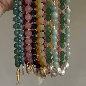May include: A collection of multi-colored beaded bracelets featuring various gemstone spheres in shades of blue, red, pink, black, yellow, and green. Each strand is accented with small gold-toned spacers and unique charms including a fish and shells.