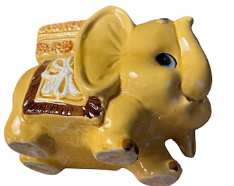 May include: A yellow ceramic elephant figurine with a brown saddle and a blue eye. The elephant is carrying a small basket on its back.