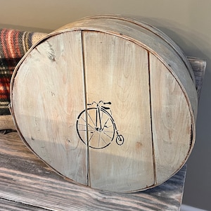 May include: A round, wooden storage container with a light gray wash finish. A black bicycle graphic is stenciled on the front. The container is sitting on a gray wooden bench.
