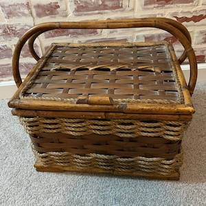 Puede incluir: Una cesta de bambú tejida marrón con un ribete de cuerda y dos asas. La cesta tiene una tapa con un patrón tejido.