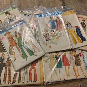 Puede incluir: Una colección de patrones de costura Simplicity vintage en fundas de plástico. Los patrones presentan ilustraciones de moda de los años 70, incluyendo vestidos, trajes de pantalón y tops. Los patrones están en varios tamaños y estilos, algunos con etiquetas de precio.
