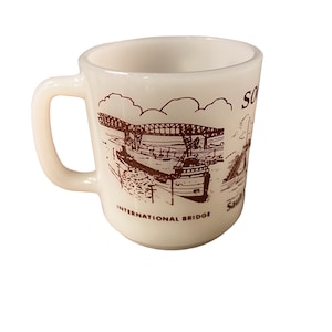 May include: White ceramic mug with a brown illustration of a bridge and a ship. The text "INTERNATIONAL BRIDGE" and "SAULT STE. MARIE" is printed on the mug.