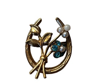 May include: Gold horseshoe pin with a bouquet of blue and white flowers. The horseshoe is decorated with a floral design and the flowers are made of rhinestones and pearls.