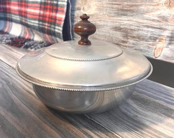 Puede incluir: Un cuenco de metal plateado con tapa. La tapa tiene un borde decorativo y un pomo de madera marrón oscuro. El cuenco está sobre una superficie de madera. Una manta a cuadros está en el fondo.