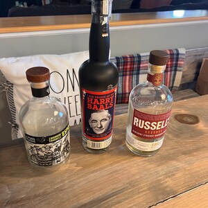 May include: Three bottles of liquor on a wooden surface. One bottle is black with a label that reads "I AM PROUD OF MY HARRY BAALS". Another bottle is clear with a label that reads "RUSSELL RESERVE". The third bottle is clear with a label that reads "PURO BOURBON BARRELS".