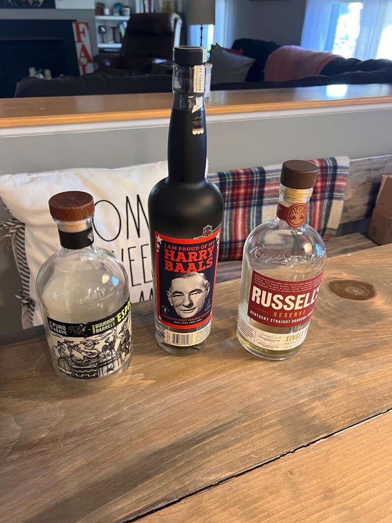 May include: Three bottles of liquor on a wooden surface. One bottle is black with a label that reads "I AM PROUD OF MY HARRY BAALS". Another bottle is clear with a label that reads "RUSSELL RESERVE". The third bottle is clear with a label that reads "PURO BOURBON BARRELS".