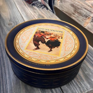 Op de afbeelding: Een ronde, marineblauwe blikken doos met een gouden rand en een decoratieve illustratie op het deksel. Het deksel heeft de tekst "GOOD HOUSEKEEPING" en een vintage afbeelding van dansende mensen. De doos is gelabeld "COMMEMORATIVE TIN".