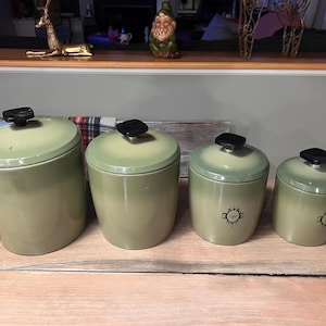 Może przedstawiać: Zestaw czterech zielonych ceramicznych pojemników kuchennych z czarnymi pokrywkami z gałkami. Pojemniki są stopniowane pod względem wielkości, a dwa najmniejsze mają czarny kwiatowy wzór i napis "Coffee". Pojemniki są ułożone na drewnianej powierzchni.