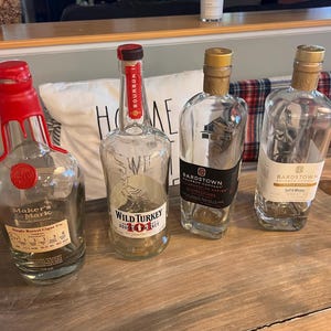 May include: Four empty bourbon bottles on a wooden surface. The bottles include Maker's Mark, Wild Turkey 101, and two Bardstown Bourbon Company bottles. The Maker's Mark bottle has a red wax seal.