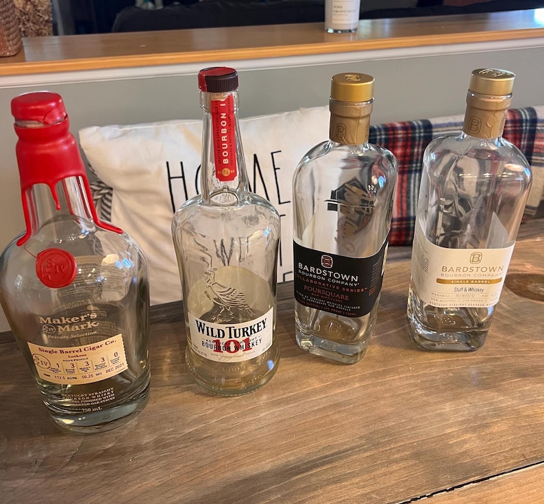 May include: Four empty bourbon bottles on a wooden surface. The bottles include Maker's Mark, Wild Turkey 101, and two Bardstown Bourbon Company bottles. The Maker's Mark bottle has a red wax seal.