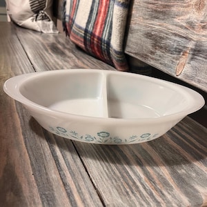 Puede incluir: Plato ovalado blanco dividido con un estampado floral turquesa. El plato tiene un acabado liso y brillante y está diseñado con dos compartimentos separados. El diseño floral se repite alrededor del borde del plato.