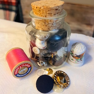 May include: A clear glass jar filled with assorted buttons, topped with a cork stopper. A pink spool of Coats & Clark's thread, a thimble, and various decorative buttons surround the jar. The thread spool reads "125 YDS. O.N.T. 50 MERCERIZED SEWING."