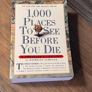 以下が含まれることがあります： パトリシア・シュルツ著の「1,000 Places To See Before You Die」というタイトルのハードカバー本。表紙は世界地図のデザインと小さな地球儀が特徴です。本のタイトルは大きく太字で書かれています。本は木製の表面にあります。
