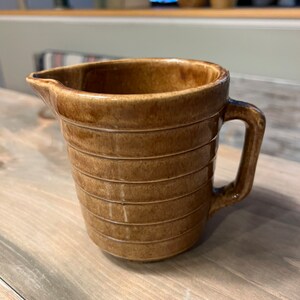 Puede incluir: Una jarra de cerámica marrón con asa y boquilla. La jarra tiene estrías horizontales alrededor de su cuerpo. La jarra está sobre una superficie de madera. En el fondo, hay otras jarras de cerámica.