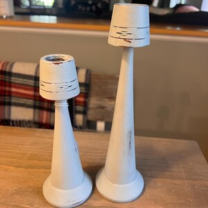 Pu&ograve; includere: Due portacandele in legno verniciato di bianco con finitura anticata. I portacandele hanno una forma conica con base rotonda e piccola sommit&agrave; cilindrica. Le sommit&agrave; presentano linee orizzontali e un aspetto leggermente invecchiato. Sono esposti su un tavolo di legno.