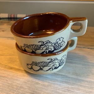 Può includere: Due ciotole per zuppa in ceramica marrone e beige con un disegno di frutta in bianco e nero. Le ciotole hanno un bordo marrone e un manico.