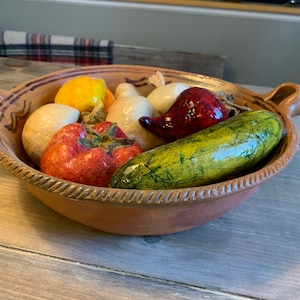 Puede incluir: Un cuenco de cerámica de color terracota, redondo, con dos asas laterales y un borde decorativo similar a una cuerda. Dentro del cuenco hay varias frutas y verduras de cerámica realistas y brillantes, incluyendo un tomate rojo y un calabacín verde.