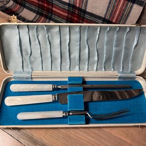 May include: An open, vintage cutlery set in a cream-colored case with a light blue satin lining. The set includes a carving knife, fork, sharpener, and two smaller utensils, all with white, pearlescent handles.