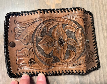 May include: A brown leather wallet with a floral design embossed on the front. The wallet has a black stitched border and a metal snap closure.