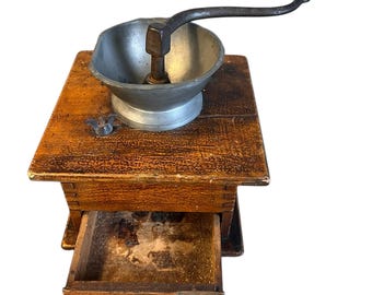 Op de afbeelding: Antiek houten koffiemolen met een metalen trechter en een lade om de koffieprut op te vangen. De molen heeft een handmatige slinger en is gemaakt van donker hout.
