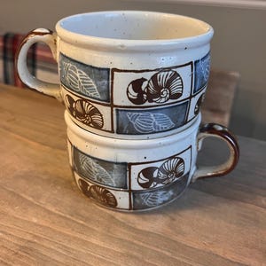 May include: Two stacked ceramic mugs with a cream-colored base and brown handles. Each mug features a repeating pattern of brown and blue squares with seashell designs. The mugs are sitting on a wooden surface.