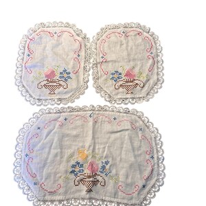 Pu&ograve; includere: Un set di tre tovagliette e sottobicchieri ricamati. Il tessuto bianco presenta un disegno a cestino floreale a punto croce rosa, blu e giallo. Ogni pezzo ha un bordo in pizzo smerlato, che aggiunge un tocco vintage.