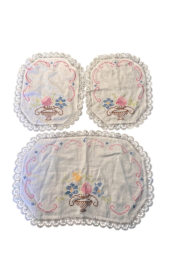 Pu&ograve; includere: Un set di tre tovagliette e sottobicchieri ricamati. Il tessuto bianco presenta un disegno a cestino floreale a punto croce rosa, blu e giallo. Ogni pezzo ha un bordo in pizzo smerlato, che aggiunge un tocco vintage.