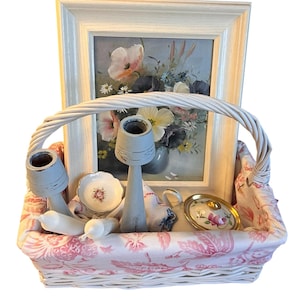 Pu&ograve; includere: Un cestino di vimini bianco foderato con tessuto floreale rosa, contenente un dipinto floreale incorniciato, due candelabri, una tazzina da t&egrave;, un piattino, un piattino e due candele bianche. Il dipinto presenta fiori rosa e gialli.