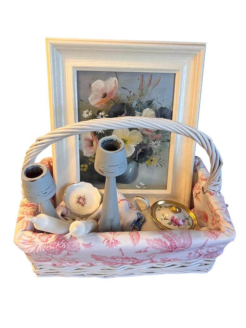 Pu&ograve; includere: Un cestino di vimini bianco foderato con tessuto floreale rosa, contenente un dipinto floreale incorniciato, due candelabri, una tazzina da t&egrave;, un piattino, un piattino e due candele bianche. Il dipinto presenta fiori rosa e gialli.