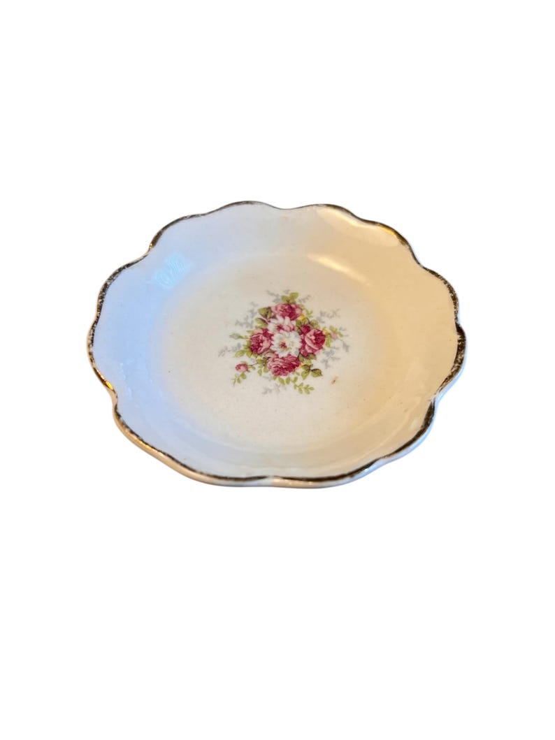 Pu&ograve; includere: Un piccolo piatto bianco con bordo smerlato e rifinitura dorata. Il centro del piatto presenta un motivo floreale con rose rosa e rosse e foglie verdi. Il piatto &egrave; probabilmente in porcellana o ceramica.