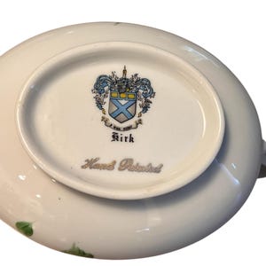 Pu&ograve; includere: Piatto in porcellana bianca con un motivo floreale e le parole "Hand Painted". Il fondo del piatto presenta uno stemma con un disegno blu e giallo e il nome "Kirk".
