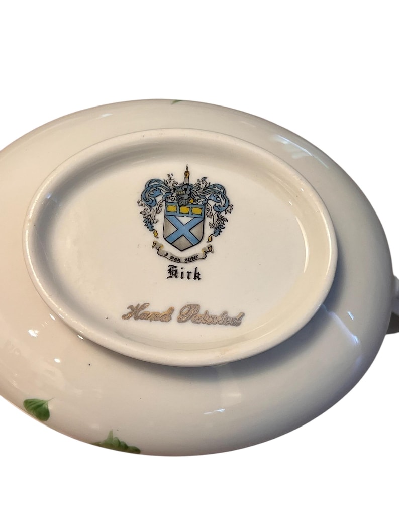 Pu&ograve; includere: Piatto in porcellana bianca con un motivo floreale e le parole "Hand Painted". Il fondo del piatto presenta uno stemma con un disegno blu e giallo e il nome "Kirk".