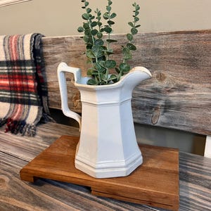 May include: A white, vintage-style pitcher with a handle, filled with green leafy stems, sits on a small, rectangular wooden tray. The pitcher has a geometric design. A plaid blanket is visible in the background.