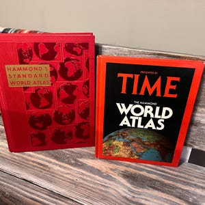 May include: Two vintage world atlases. The red atlas on the left is titled "Hammond's Standard World Atlas." The atlas on the right is titled "Time The Hammond World Atlas." Both books are on a wooden surface.