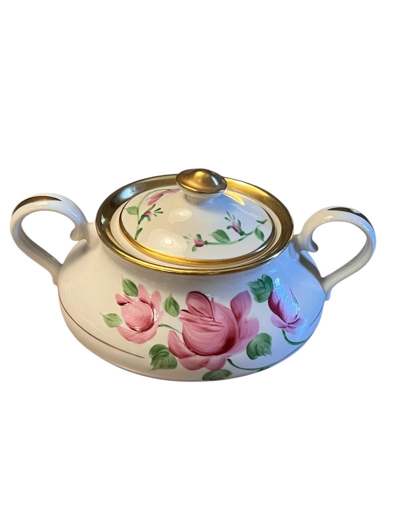 Pu&ograve; includere: Zuccheriera in ceramica bianca con coperchio, con rose rosa e foglie verdi dipinte a mano. I bordi e le maniglie sono dorati. La ciotola ha due manici e un pomello dorato sul coperchio.