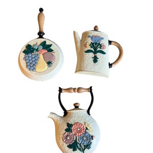 May include: Set of three white ceramic wall decor pieces in the shape of kitchen items. The pieces are a teapot, a coffee pot, and a frying pan. Each piece has a floral or fruit design.
