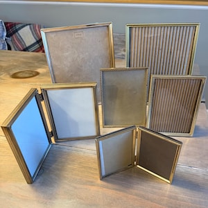 Puede incluir: Una colección de seis marcos de fotos de metal dorado con bordes finos y perlados. El conjunto incluye dos marcos dobles con bisagras y cuatro marcos individuales de varios tamaños, algunos con inserciones lisas y otros con papel acanalado.