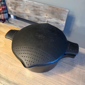 May include: A black Pampered Chef cooking pot with a matching lid featuring integrated straining holes on two sides. The lid has two side handles and the brand name embossed in the center, while the pot body is round and solid black.
