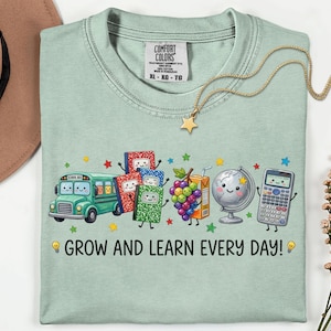 May include: A light green t-shirt featuring a graphic of a smiling school bus, colorful notebooks, a bunch of grapes, a juice box, a globe, and a calculator. The text "GROW AND LEARN EVERY DAY!" is printed below the characters with lightbulb icons.