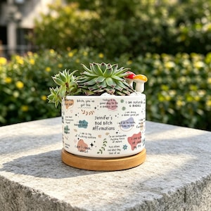 May include: A white ceramic cylindrical planter with a bamboo saucer, featuring colorful text and abstract shapes. The text reads Jennifer's Bad Bitch Affirmations along with various phrases. A green succulent and two small mushroom ornaments are inside.