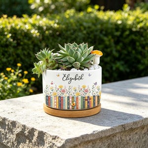 May include: A white ceramic planter with a wooden base containing a green succulent and a small mushroom figurine. The planter features a printed design of colorful books, wildflowers, and butterflies, with the name Elizabeth in black script.