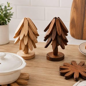 May include: Two wooden tree-shaped structures composed of stacked, leaf-like slats, one in light wood and one in dark wood. A matching dark wood circular coaster set is laid flat, and another light wood coaster set sits beneath a white ceramic pot.