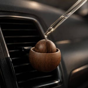 May include: A spherical dark brown wooden car air freshener diffuser attached to a black plastic vehicle air vent. A clear glass dropper with a black rubber bulb is positioned above the wooden sphere, releasing a single drop of liquid.
