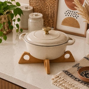 May include: A cream-colored enameled cast iron Dutch oven with a matching lid and a gold-toned knob, resting on a light-colored wooden trivet. The pot features two side handles and a smooth, speckled finish, providing a functional kitchen vessel.