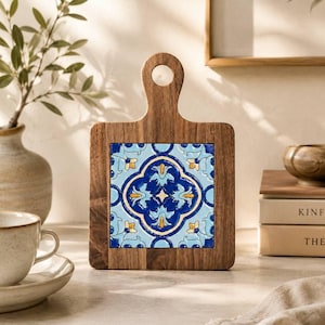 May include: A square wooden serving board with a handle featuring a central ceramic tile inlay. The tile displays a symmetrical floral pattern in shades of deep blue, light blue, and yellow, set against a dark brown wood grain frame.