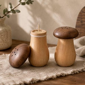 May include: Two mushroom-shaped wooden containers with dark brown caps and light tan bases. One container is open, revealing a collection of wooden toothpicks inside, while the other is closed. The caps feature small holes on the top surface.