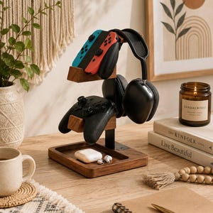 May include: A multi-tiered wooden gaming accessory stand featuring a rectangular base tray, a central vertical support, and two horizontal arms. The stand holds a red and blue game controller, a black game controller, and black over-ear headphones.