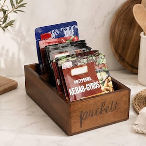 May include: A rectangular dark brown wooden storage box with the word packets engraved on the front side. The box contains several upright food seasoning packets, including one clearly labeled PRZYPRAWA KEBAB-GYROS with a graphic of food.