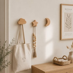 May include: Three light-colored wooden wall hooks shaped like a cloud, a star, and a crescent moon. A beige canvas tote bag hangs from the cloud hook, and a decorative string of wooden beads with a tassel hangs from the star-shaped hook.